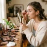 Woman applying makeup at white vanity with scattered beauty products in soft morning light