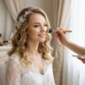 Bride receiving professional makeup application near window with natural light and elegant preparation atmosphere