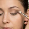 Woman holding fluffy blending brush with proper grip while applying neutral eyeshadow in natural lighting