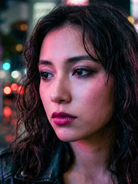 Woman with makeup on city street at night with colorful neon lighting creating pink and blue reflections
