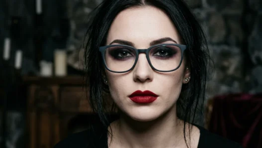 Woman wearing glasses with dramatic vampire makeup showing dark eyeshadow and deep red lips in gothic lighting