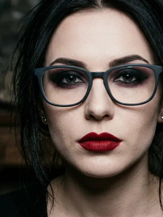 Woman wearing glasses with dramatic vampire makeup showing dark eyeshadow and deep red lips in gothic lighting