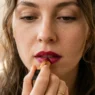 Woman applying bold lipstick by hand mid-swipe across lower lip in soft warm lighting
