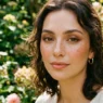 Woman with scattered warm auburn faux freckles and peachy blush in a lush green garden with soft bokeh light