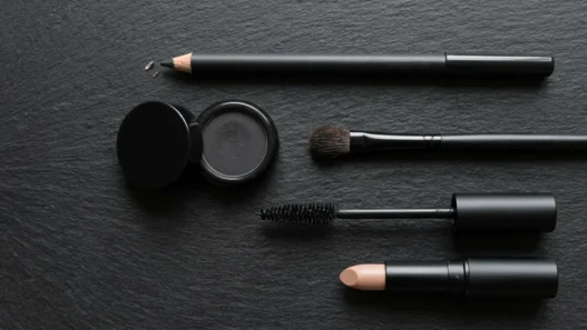 Flat lay of emo makeup essentials including kohl pencil, black eyeshadow, brush, mascara and nude lip on black surface