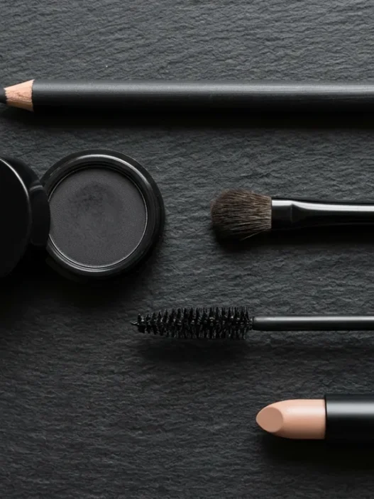 Flat lay of emo makeup essentials including kohl pencil, black eyeshadow, brush, mascara and nude lip on black surface