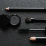 Flat lay of emo makeup essentials including kohl pencil, black eyeshadow, brush, mascara and nude lip on black surface