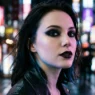 Woman with jet black smoky eye and near-black plum lipstick standing on a city street under neon light at night
