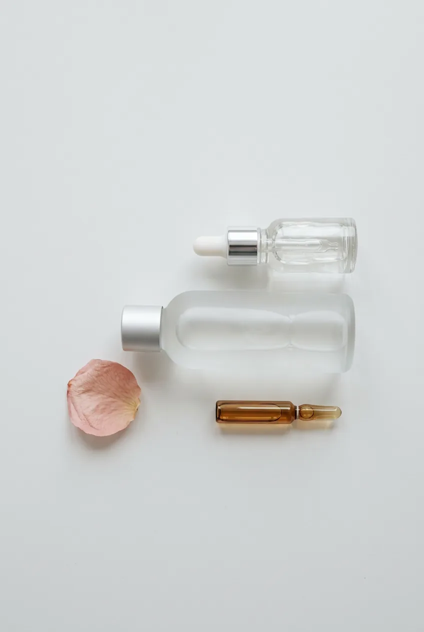 Hydrating toner bottle, serum dropper, and ampoule arranged minimally on a white matte surface with a dried petal
