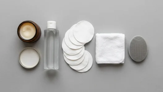 Flat lay of makeup removal essentials including cleansing balm, micellar water, and reusable cotton rounds on a grey matte surface