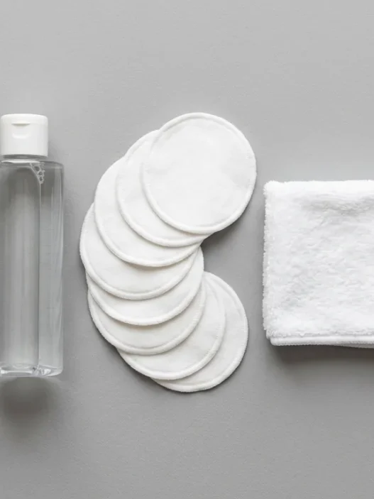 Flat lay of makeup removal essentials including cleansing balm, micellar water, and reusable cotton rounds on a grey matte surface