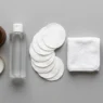 Flat lay of makeup removal essentials including cleansing balm, micellar water, and reusable cotton rounds on a grey matte surface