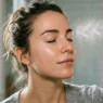 Woman's face with fine mist of facial spray above cheekbone in soft natural window light, skin texture visible