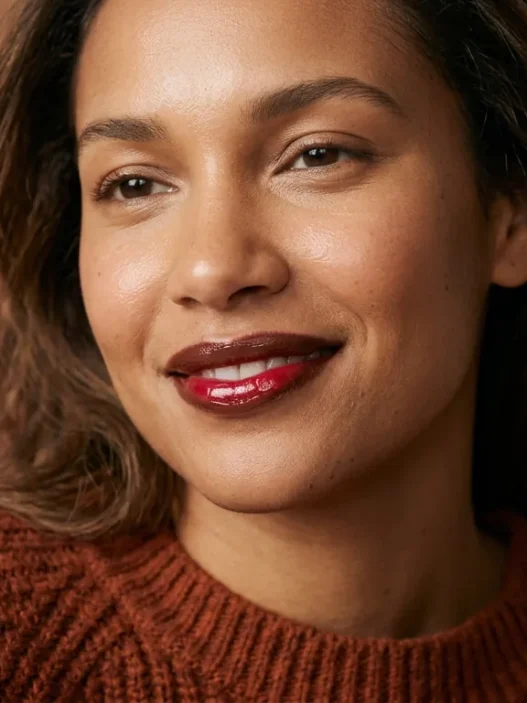 Woman wearing glossy cocoa and cherry lip combo in rust sweater under soft studio light