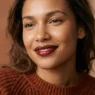 Woman wearing glossy cocoa and cherry lip combo in rust sweater under soft studio light