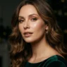 Woman with chestnut waves wearing glowing neutral makeup with nude gloss against dark background